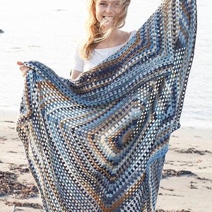 Puede incluir: Una manta de ganchillo grande y cuadrada con un patrón geométrico en tonos azul marino, azul claro, gris y beige. El tejido está construido con un diseño de punto granny square calado, creando una apariencia texturizada y ligera.