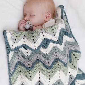Puede incluir: Una manta de bebé tejida a ganchillo con un patrón de chevrón en rayas alternas de color verde salvia, azul pizarra, gris claro y blanco. La manta tiene un diseño de punto calado y cubre a un bebé dormido que usa un chupete gris.