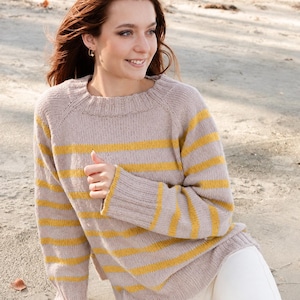Puede incluir: Un suéter de punto de manga larga con una base de color beige claro y rayas horizontales de color amarillo mostaza. La prenda presenta un cuello redondo, puños acanalados y un dobladillo acanalado, confeccionado con un patrón de punto liso visible.