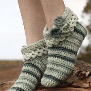 Puede incluir: Un par de zapatillas de ganchillo a la altura del tobillo con rayas horizontales en varios tonos de verde salvia y verde bosque oscuro. Las zapatillas tienen un borde festoneado decorativo y dos pequeñas flores de ganchillo superpuestas.