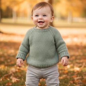 Puede incluir: Un bebé sonriente con un suéter de punto verde salvia y pantalones grises claros está de pie sobre hierba con hojas caídas. El bebé también lleva zapatos de cuero marrón. El fondo borroso tiene colores otoñales.