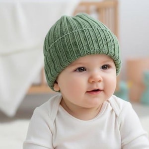Puede incluir: Un bebé con un gorro de punto acanalado verde y un body blanco. El bebé mira hacia el lado derecho con una ligera sonrisa. El fondo incluye una cuna y una manta suave.
