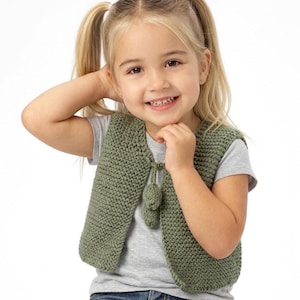 Puede incluir: Una niña pequeña con un chaleco verde tejido con cierre de lazo. El chaleco es de color liso y parece hecho a mano. La niña lleva una camiseta gris y vaqueros azules. El fondo es blanco.
