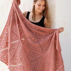 Puede incluir: Una manta grande de punto en color rosa empolvado que presenta un intrincado patrón de encaje con formas geométricas, incluyendo diamantes y triángulos, creados mediante puntos calados e hilo texturizado, con un diseño cuadrado.