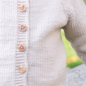 Puede incluir: Una chaqueta de punto color crema con cuello acanalado y una tapeta vertical con seis botones de madera de color marrón claro, cada uno grabado con un pequeño corazón. El texto GARN STUDIO DROPS DESIGN www.garnstudio.com es visible.