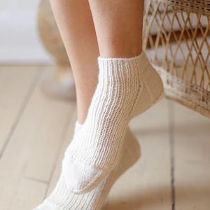 Puede incluir: Un par de calcetines tobilleros de color crema claro. Los calcetines presentan un patrón de punto acanalado vertical en toda la parte principal, con una textura de punto lisa y sólida en las secciones del talón y la puntera. El tejido es suave.