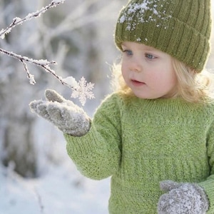 Könnte beinhalten: Ein Kind mit einer grünen Strickmütze, einem Pullover und Fäustlingen, bedeckt mit Schnee. Das Kind betrachtet eine Schneeflocke auf seinem Fäustling. Der Hintergrund ist eine verschwommene, verschneite Winterszene.