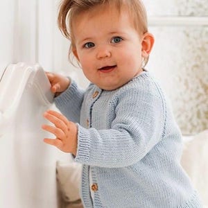 Puede incluir: Una chaqueta de punto de manga larga en color azul claro con cuello redondo, puños y dobladillo acanalados. La prenda se abrocha en la parte delantera con cuatro pequeños botones redondos de madera de color marrón claro, de diseño sencillo.
