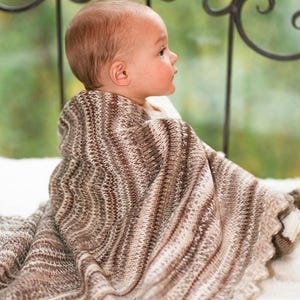 Puede incluir: Una manta de bebé tejida suave con un patrón variado de rayas marrones, bronceadas y crema. La manta tiene un diseño de punto ondulado texturizado y un borde festoneado, drapeada sobre los hombros de un niño pequeño con cabello castaño claro.