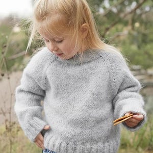 Puede incluir: Un suéter de punto gris claro con cuello redondo y mangas largas. El suéter lo lleva un niño pequeño, combinado con pantalones cortos de mezclilla azules. El niño sostiene dos galletas pequeñas y rectangulares. El fondo está borroso, lo que sugiere un entorno al aire libre.