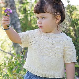 Puede incluir: Una blusa de punto color crema de manga corta con un delicado patrón de encaje festoneado en el canesú y el pecho, combinada con pantalones de mezclilla azul. La prenda presenta un cuello redondo y un dobladillo acanalado con textura.
