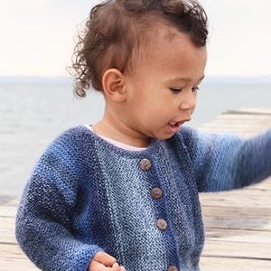 Puede incluir: Un cárdigan de punto de manga larga con un patrón de color degradado que transiciona de azul marino oscuro a tonos más claros de azul y blanco. La prenda tiene un cuello redondo y se abrocha en la parte delantera con cuatro botones marrones.