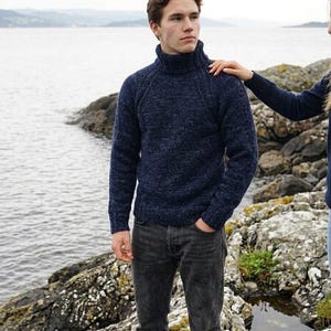 Puede incluir: Un jersey de cuello alto azul marino con un diseño de punto texturizado. El jersey se combina con vaqueros grises oscuros. La persona está de pie en una costa rocosa con el océano al fondo.