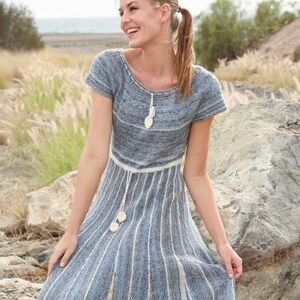 Puede incluir: Un vestido de punto de manga corta en tonos azules y grises, con cuello redondo, un cordón decorativo blanco con pompones en la cintura y una falda acampanada con rayas verticales blancas que crean una apariencia acanalada y texturizada.