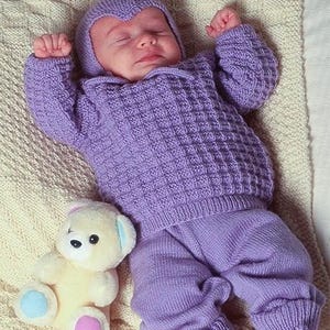 Puede incluir: Un conjunto de punto para bebé en color lavanda que consta de un suéter de manga larga con patrón de textura de gofre, pantalones a juego con puños acanalados y una capucha puntiaguda. Un pequeño oso de peluche color crema con patas azules y rosas está cerca.