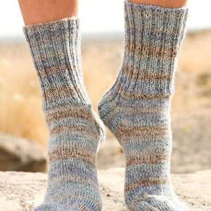 Puede incluir: Un par de calcetines tejidos a mano en tonos azules, marrones y grises. Los calcetines tienen un puño acanalado y un patrón de rayas. Los calcetines se llevan en los pies.