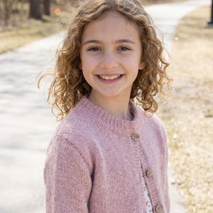 Puede incluir: Una persona con cabello rizado sonríe, usando un cárdigan rosa claro con botones de madera. El cárdigan está abrochado y la persona está afuera en un día soleado, con un camino en el fondo.