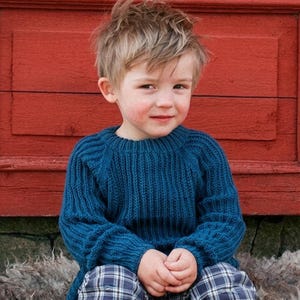 May include: A child wearing a deep teal blue long-sleeved jumper with a ribbed knit texture and a crew neckline, paired with blue and white plaid trousers. The jumper features raglan sleeves and a thick ribbed collar, cuffs, and hemline.