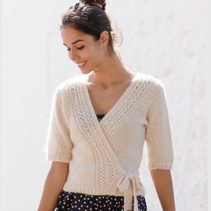 Puede incluir: Un suéter cruzado de punto color crema con mangas cortas, cuello en V y cierre de lazo lateral. La prenda presenta un patrón de trenzas texturizado a lo largo de los bordes delanteros y puños acanalados, combinado con una falda oscura con lunares blancos.