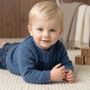 Puede incluir: Un niño pequeño con un suéter de punto azul y jeans está acostado sobre una alfombra color crema. El niño tiene cabello rubio y ojos azules. Bloques de letras de madera en el fondo. Una escena acogedora.