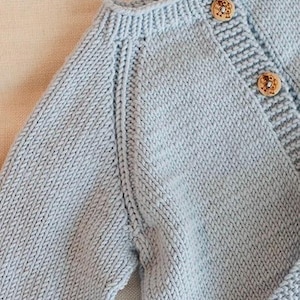Puede incluir: Un cárdigan de punto azul claro con cuello redondo acanalado, mangas largas con costuras raglán y una tapeta de botones frontal. La prenda se cierra con dos botones redondos de madera marrón claro con un diseño floral tallado.