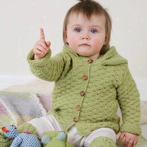 Puede incluir: Una chaqueta de punto de color verde claro con capucha y cinco botones marrones en la parte delantera. La prenda presenta un patrón de punto texturizado en el cuerpo y las mangas. La chaqueta se combina con calentadores de punto a juego.