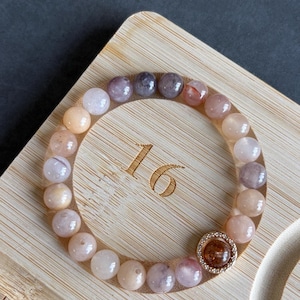 May include: A beaded bracelet featuring a variety of pastel-colored gemstones, including pink, purple, and peach tones. The bracelet has a gold-colored accent piece and is displayed on a wooden surface. The beads are approximately 0.3 inches in diameter.