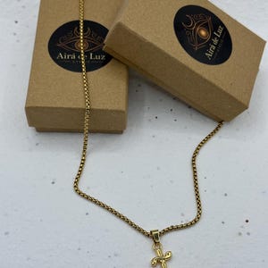 Könnte beinhalten: Eine goldfarbene Halskette mit einer Box-Kette und einem dreizackförmigen Anhänger. Die Kette wird zusammen mit zwei braunen Pappschachteln präsentiert, die jeweils einen runden schwarzen Aufkleber mit dem Text Airá de Luz Atelie tragen.
