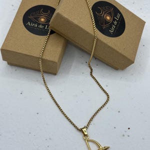 May include: A gold-toned box chain necklace featuring a pendant shaped like a bow and arrow. The necklace is displayed alongside two brown cardboard gift boxes, each featuring a circular black sticker with the text Airá de Luz ATELIÊ.