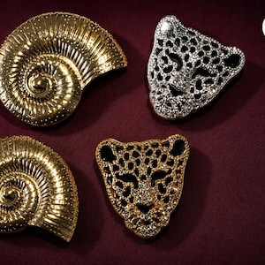 May include: Two gold-toned metallic ammonite shell brooches with ribbed textures and two leopard head brooches, one in silver-tone and one in gold-tone, both featuring intricate cut-out patterns and embedded clear crystals throughout the designs.