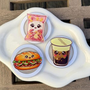 May include: Three circular phone grips featuring kawaii-style illustrations of food items: a pink bag of shrimp chips, a Vietnamese banh mi sandwich with fresh vegetables, and a cup of brown sugar bubble tea with a creamy yellow foam topping.