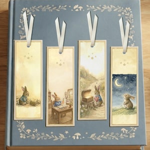 May include: Four rectangular paper bookmarks with cream-colored backgrounds and white ribbon tassels. Each features a watercolor illustration of a rabbit in different settings, including reading, studying, holding a heart, and looking at a crescent moon.
