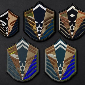 May include: Five shield-shaped embroidered patches featuring the United States Air Force enlisted rank insignia. Each patch displays a central star, chevron stripes, and a combination of black, tan, brown, blue, and camouflage fabric patterns.