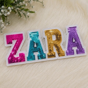 Puede incluir: Un parche decorativo con el nombre ZARA en letras de bloque. Cada letra está cubierta de lentejuelas: la Z es rosa fuerte, la A es turquesa, la R es dorada y la última A es morada. Cada letra tiene un borde de fieltro blanco que la perfila.