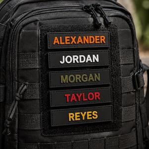 May include: A black tactical backpack featuring five rectangular hook-and-loop patches attached to a central panel. The patches display the names ALEXANDER in orange, JORDAN in white, MORGAN in olive green, TAYLOR in red, and REYES in yellow.