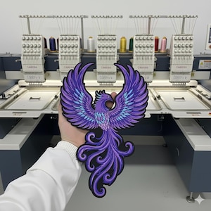 Puede incluir: Un parche bordado que muestra un fénix con las alas extendidas hacia arriba. El diseño utiliza hilos en tonos morado, violeta y azul claro con un borde negro. El fénix tiene una cola larga y fluida con patrones intrincados y arremolinados.