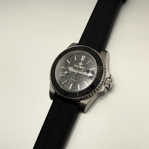 May include: A stainless steel wristwatch with a black bezel and a black rubber strap. The watch face is dark gray with the word "CALDERA" and the word "AUTOMATIC" printed on it. The watch is angled diagonally across the frame.