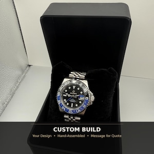 May include: A stainless steel wristwatch with a black dial and blue bezel, displayed in a black box. The watch features a silver band and the text "CUSTOM BUILD". The watch has a date display and luminous hands.