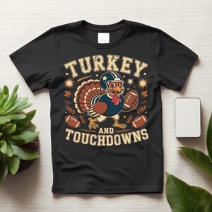 May include: A black short-sleeved t-shirt featuring a graphic of a turkey wearing a football helmet and holding a football. The text reads TURKEY AND TOUCHDOWNS. The design includes autumn leaves and stars in shades of orange, tan, and blue.