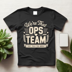 May include: A black short-sleeved t-shirt featuring cream-colored text that reads We're That OPS TEAM THEY TOLD YOU ABOUT, accented with a small heart, stars, and leaf-like flourishes. The design is centered on the chest of the garment.