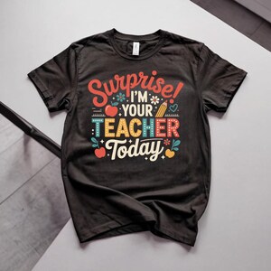 Può includere: Una t-shirt nera a maniche corte con una stampa grafica colorata che riporta il testo "Surprise! I'M YOUR TEACHER Today". Il design include illustrazioni di mele rosse, una matita gialla, piccoli fiori e cuori nei colori rosso, giallo e verde acqua.