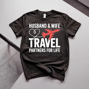 May include: A black short-sleeve t-shirt featuring white text that reads HUSBAND & WIFE, TRAVEL, and PARTNERS FOR LIFE. A dashed white heart outline and a red airplane graphic are positioned between the words HUSBAND & WIFE and TRAVEL.