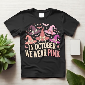 May include: A black short-sleeve t-shirt featuring a graphic print of five witch hats in shades of pink and purple, accented with small bats, stars, and hearts. The text "IN OCTOBER WE WEAR PINK" is printed in large, bold, light-colored lettering.