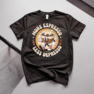 Puede incluir: Una camiseta negra de manga corta con un gráfico de una taza de café de dibujos animados con brazos y piernas caminando. El diseño incluye el texto MORE ESPRESSO LESS DEPRESSO en un marco circular beige con estrellas y granos de café.