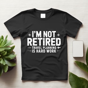 Puede incluir: Una camiseta negra de manga corta con texto blanco de estilo desgastado que dice I'M NOT RETIRED TRAVEL PLANNING IS HARD WORK, acompañada de pequeños iconos de estrellas y un gráfico de un avión en la parte frontal.