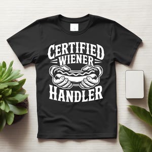 May include: A black short-sleeved t-shirt featuring a white graphic print of two hands holding a hot dog. The text above the graphic reads CERTIFIED and WIENER, while the text below the graphic reads HANDLER in a bold, stylised white font.