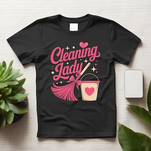 以下が含まれることがあります： ピンクの筆記体で「Cleaning Lady」という文字がプリントされた黒の半袖Tシャツ。デザインには、リボンのついたピンクのほうき、ピンクのハートが描かれたベージュのバケツ、そして周囲に小さなピンクの星とハートが配置されています。