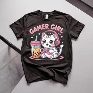 May include: A black short-sleeve t-shirt featuring a graphic of a white cat wearing pink headphones and holding a pink game controller. Next to the cat is a cup of bubble tea. The text GAMER GIRL is printed in pink above the cat illustration.