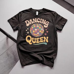 Könnte beinhalten: Ein schwarzes Kurzarm-T-Shirt mit einem Grafikdruck einer bunten Discokugel in der Mitte. Der Text DANCING QUEEN ist in einer geschwungenen Retro-Schriftart über und unter der Discokugel gedruckt, akzentuiert durch kleine Sterne und Herzen.