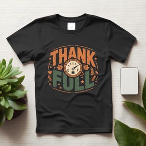 Puede incluir: Una camiseta negra de manga corta con un diseño gráfico que incluye el texto THANK FULL. El diseño presenta un icono de indicador de combustible que señala lleno, rodeado de hojas de otoño naranjas y verdes, espigas y estrellas.